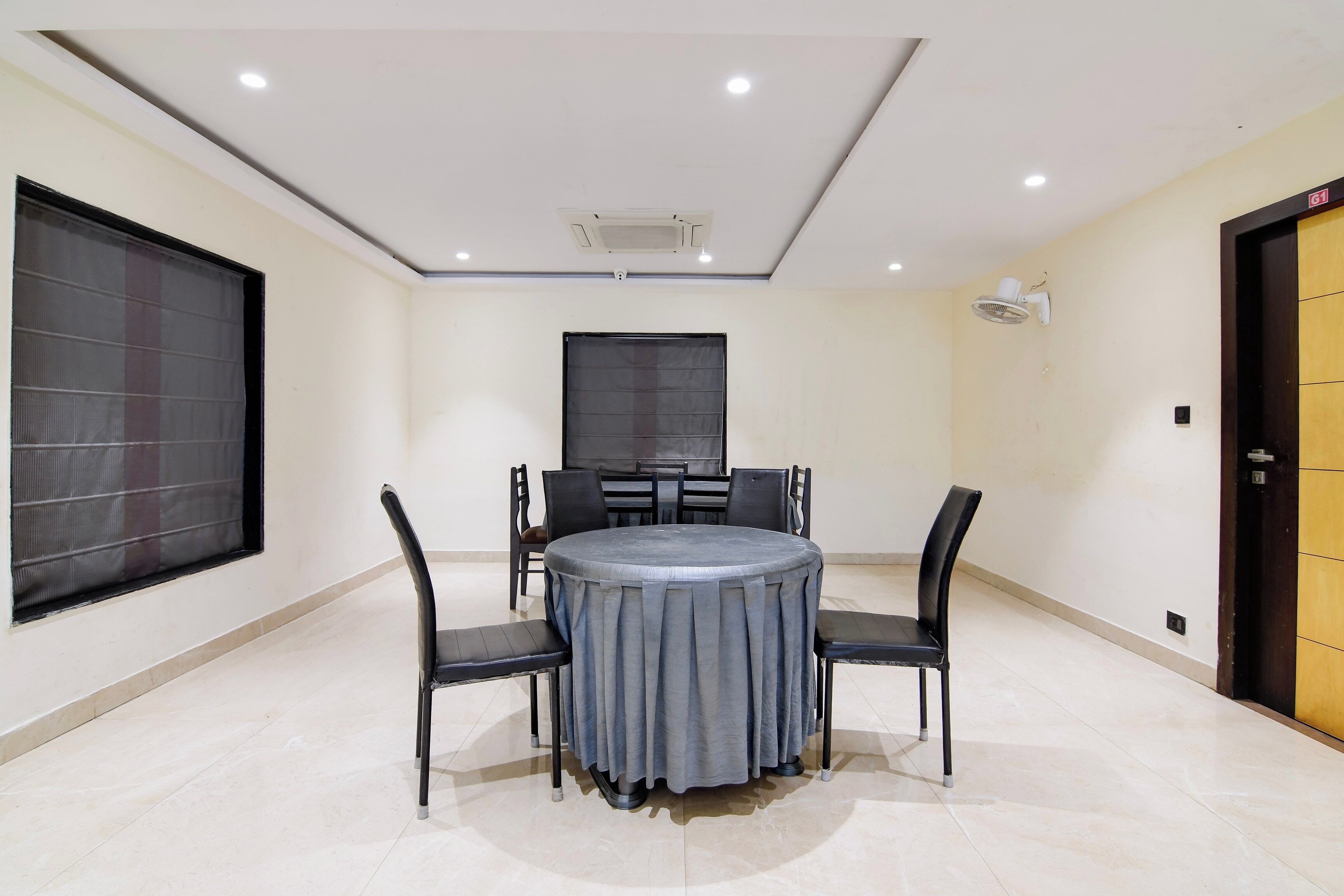 A conference room with a round table, four chairs, a large window with blinds, a door, and a ceiling fan.