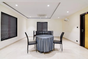 Un salón de conferencias con una mesa redonda, cuatro sillas, un amplio ventanal con persianas, una puerta y un ventilador de techo.