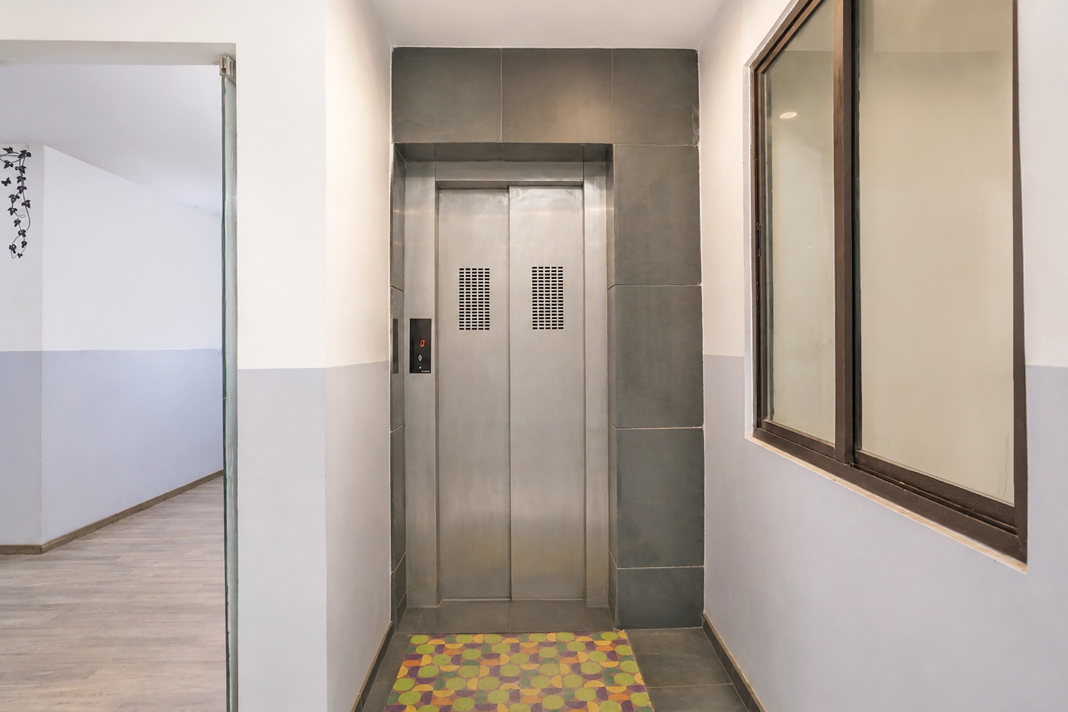 A hallway with a modern elevator, a patterned rug, and a glass door on the left.