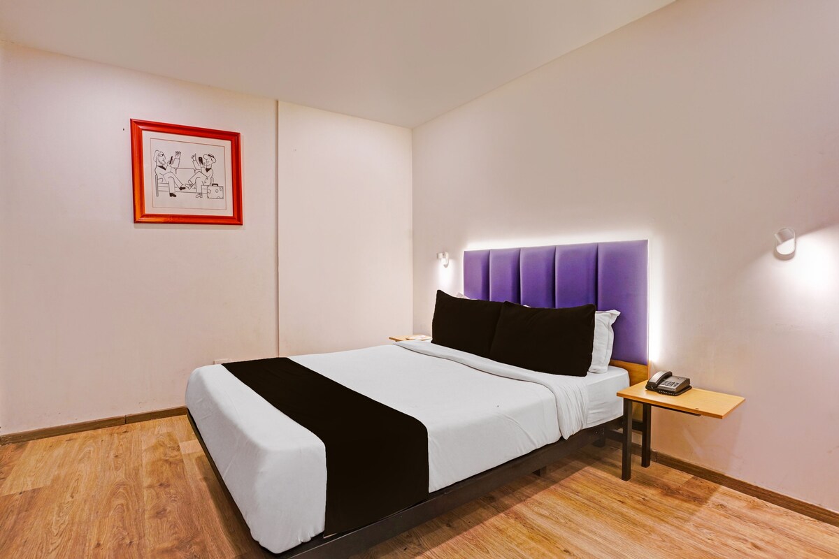 A hotel room with a bed, a nightstand, a telephone, a framed picture on the wall, and a light fixture above the bed.
