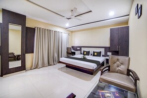 Quarto de hotel com cama, poltrona, mesa, espelho, cortinas e ventilador de teto.