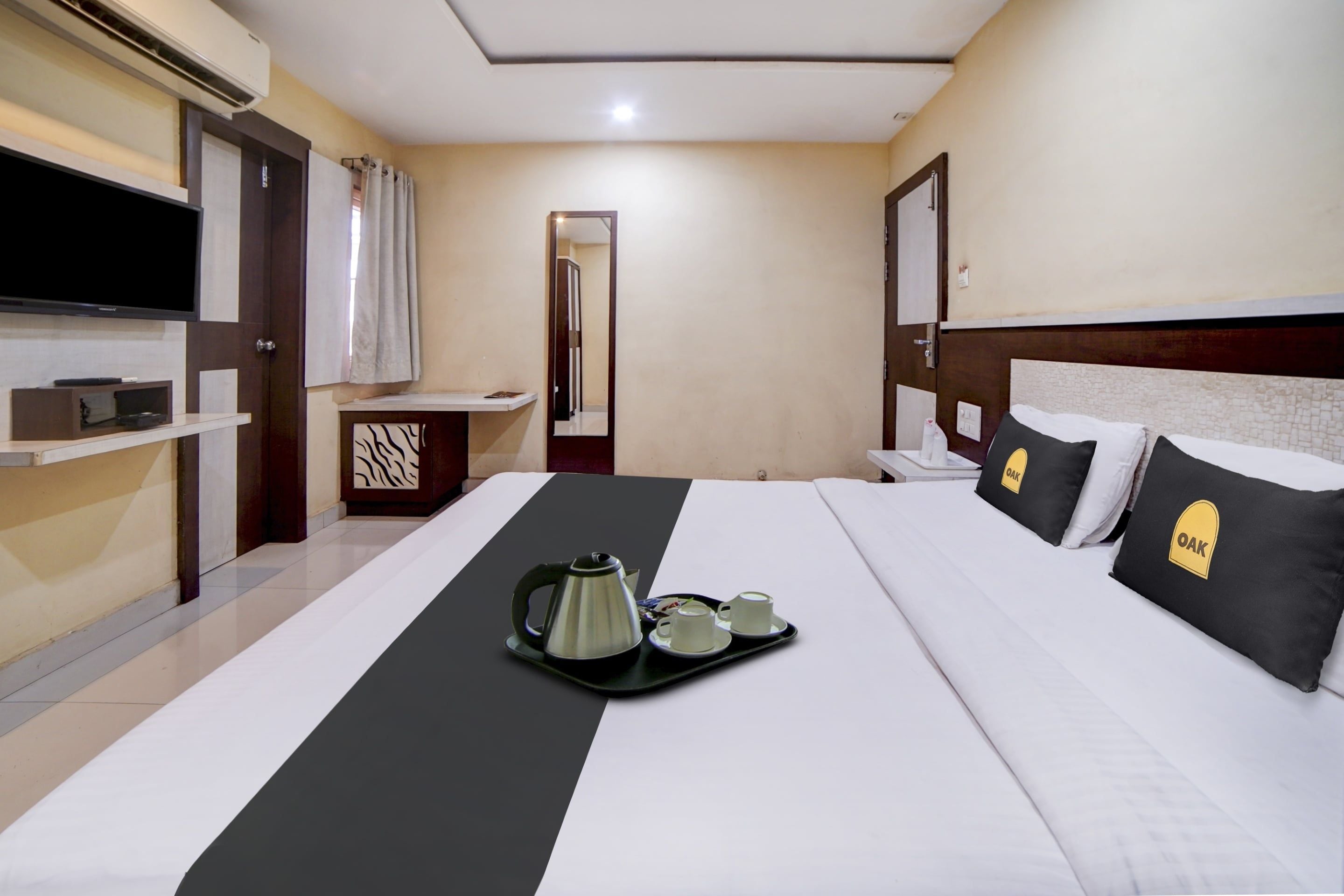 Habitación de hotel con cama, una bandeja con hervidor y tazas, televisor y espejo.