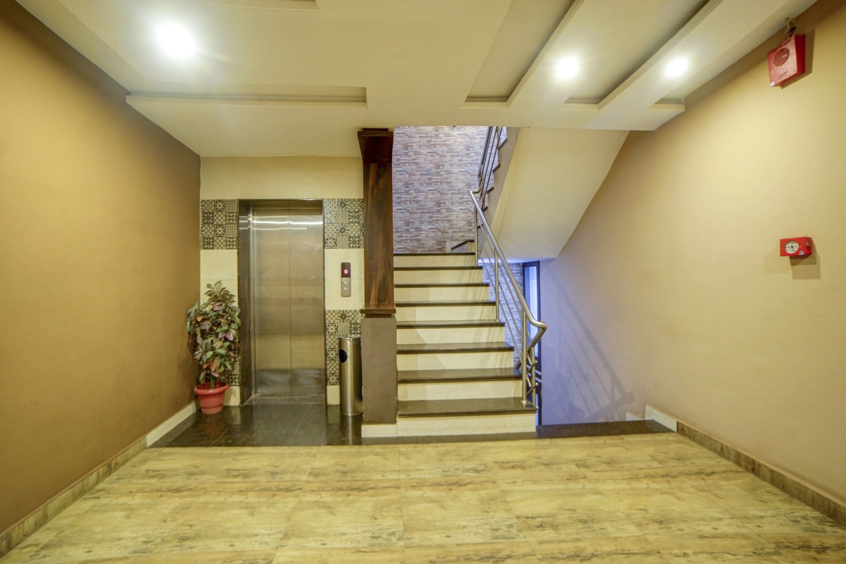 Un pasillo de hotel con suelo de mármol, una planta en maceta, un ascensor y una escalera con barandilla de metal.