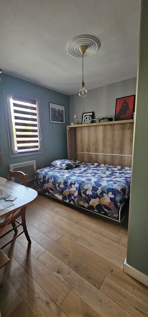 Een kamer met een bed, een tafel, een stoel, een raam met jaloezieën, een plafondlamp en een muur met een ingelijst schilderij.