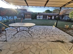 Een terras met een ronde tafel en stoelen, een overdekt gedeelte en uitzicht op een achtertuin met een huis en een fiets.
