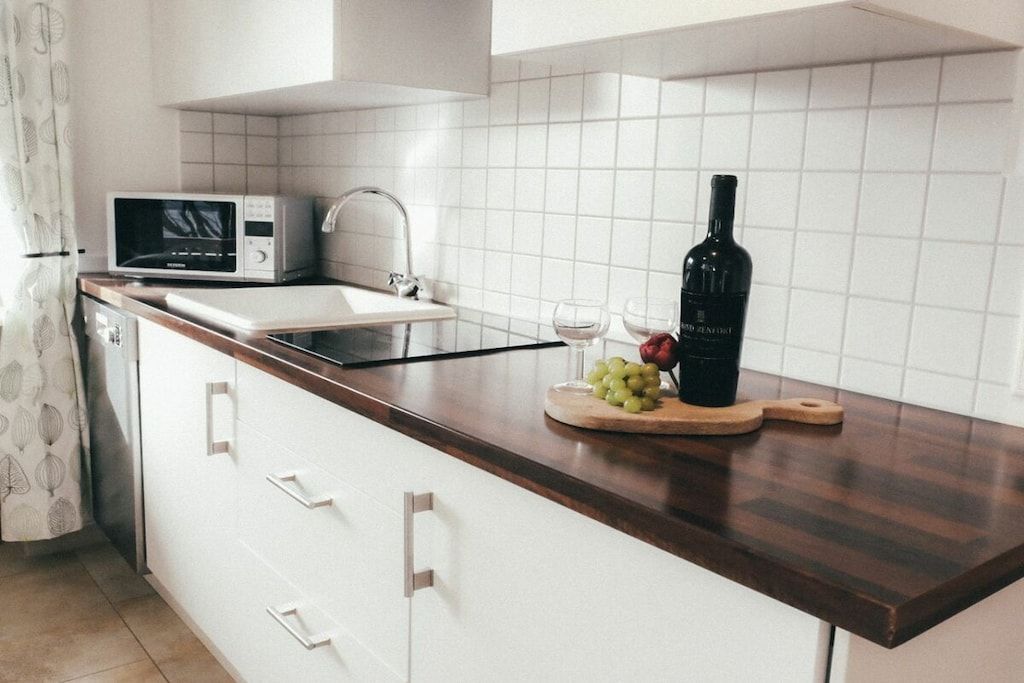 A kitchen with a microwave, sink, and a wooden cutting board with grapes and a bottle.