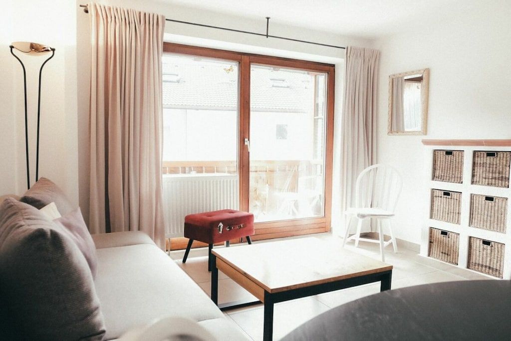 A modern living room with a sofa, a red ottoman, a white chair, a wooden table, and a large sliding glass door leading to a balcony.