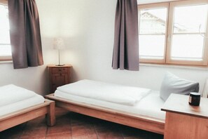 Une chambre avec deux lits simples, une petite table de chevet en bois, une lampe et une fenêtre avec des rideaux.