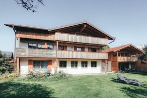 Ein zweistöckiges Gebäude mit Balkon, überdachter Terrasse und einem Rasenstück mit Liegestühlen.