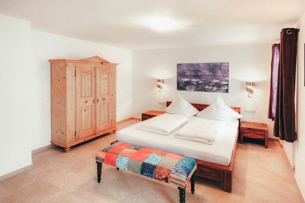 A bedroom with a bed, a wooden wardrobe, a bench, two bedside tables, a wall-mounted lamp, and a painting.