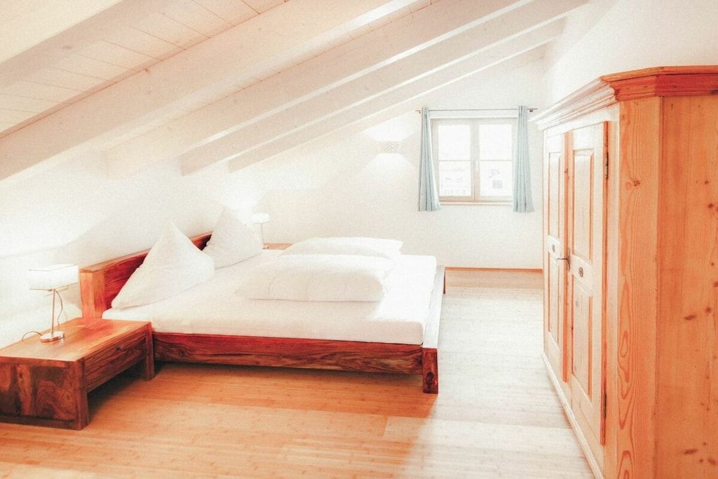 Ein Schlafzimmer mit schrägem Dach, einem Bett, einem Nachttisch, einem Kleiderschrank und einem Fenster mit Vorhängen.
