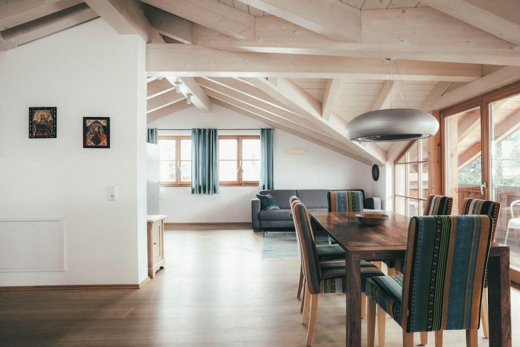 Ein Essbereich mit Holztisch und Stühlen, eine Couch im Hintergrund und ein großes Fenster mit Vorhängen.