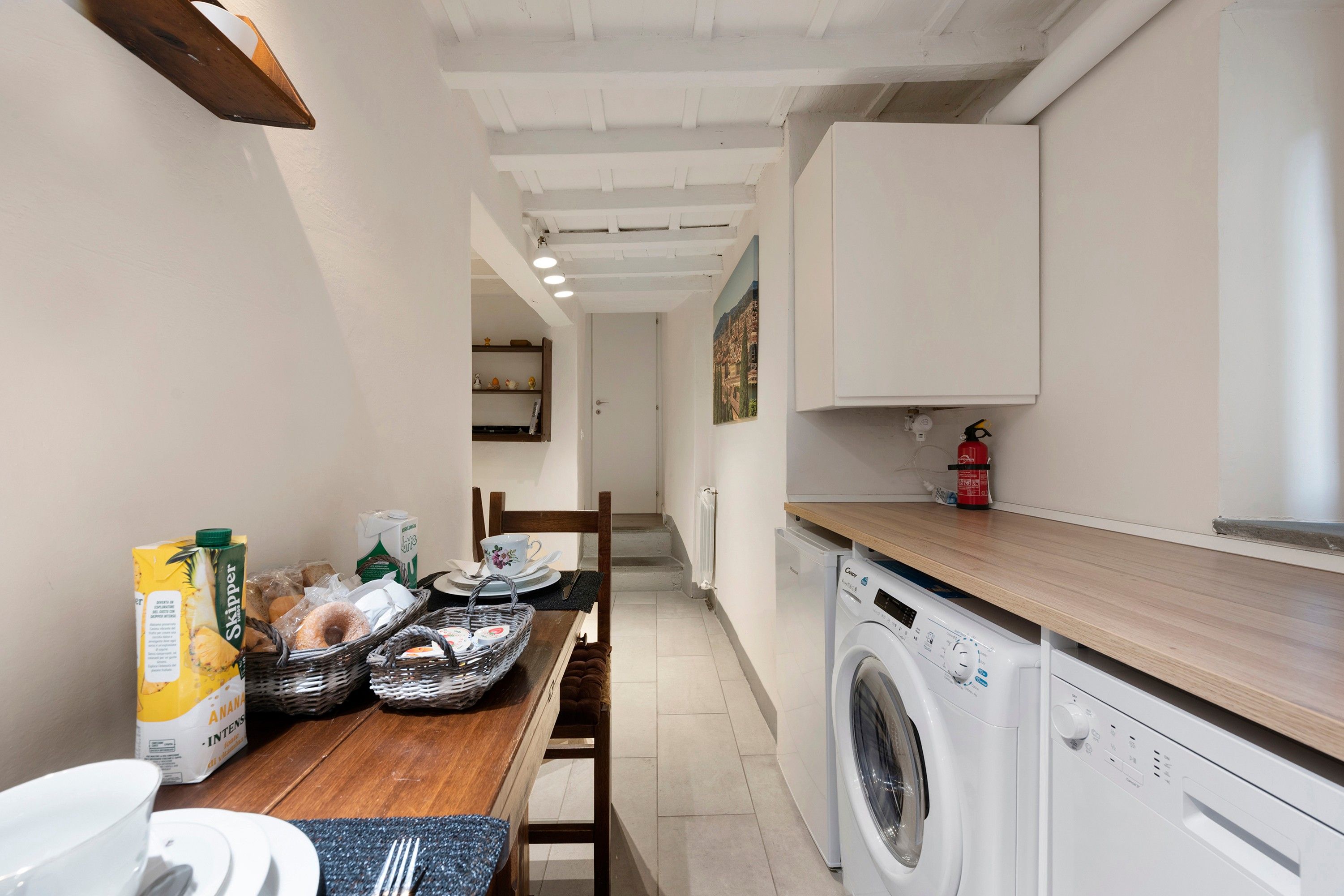 Une cuisine compacte équipée d’un lave-linge, d’une table à manger avec des aliments pour le petit-déjeuner, et offrant une vue sur le paysage urbain par une fenêtre.