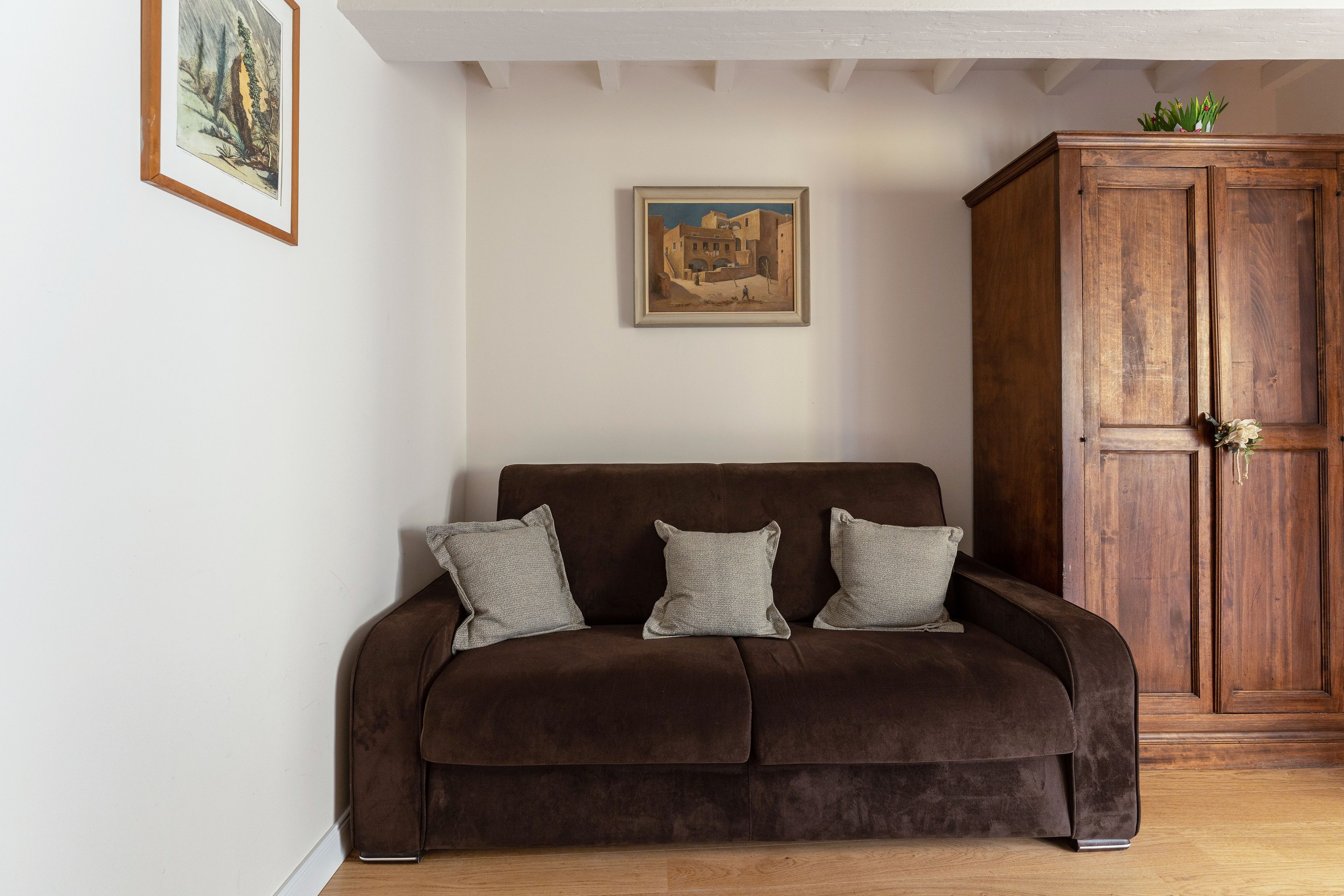 A brown sofa with three pillows against a white wall, a wooden cabinet, and a framed painting above the sofa.
