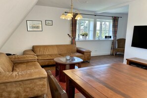 A living room with a sectional sofa, a wooden dining table, a small round table, and a television mounted on the wall. - Holiday apartment in Sylt with swimming pool (Sylt)