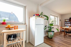 A compact kitchen with a white refrigerator, a wooden table with a toaster, a potted plant, and a window with a view of a house. - Uthland on the beach (Sylt)