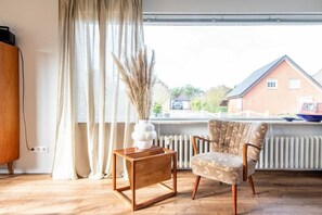 A room with a wooden floor, a chair, a small table, a radiator, and a window with curtains. - Uthland on the beach (Sylt)