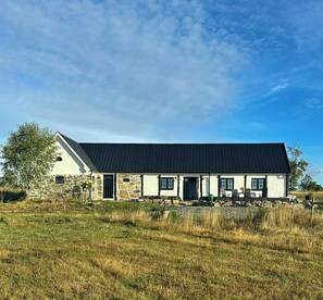 Ein zweistöckiges Steinhaus mit schwarzem Dach, einer Veranda und einem grasbewachsenen Feld im Vordergrund.