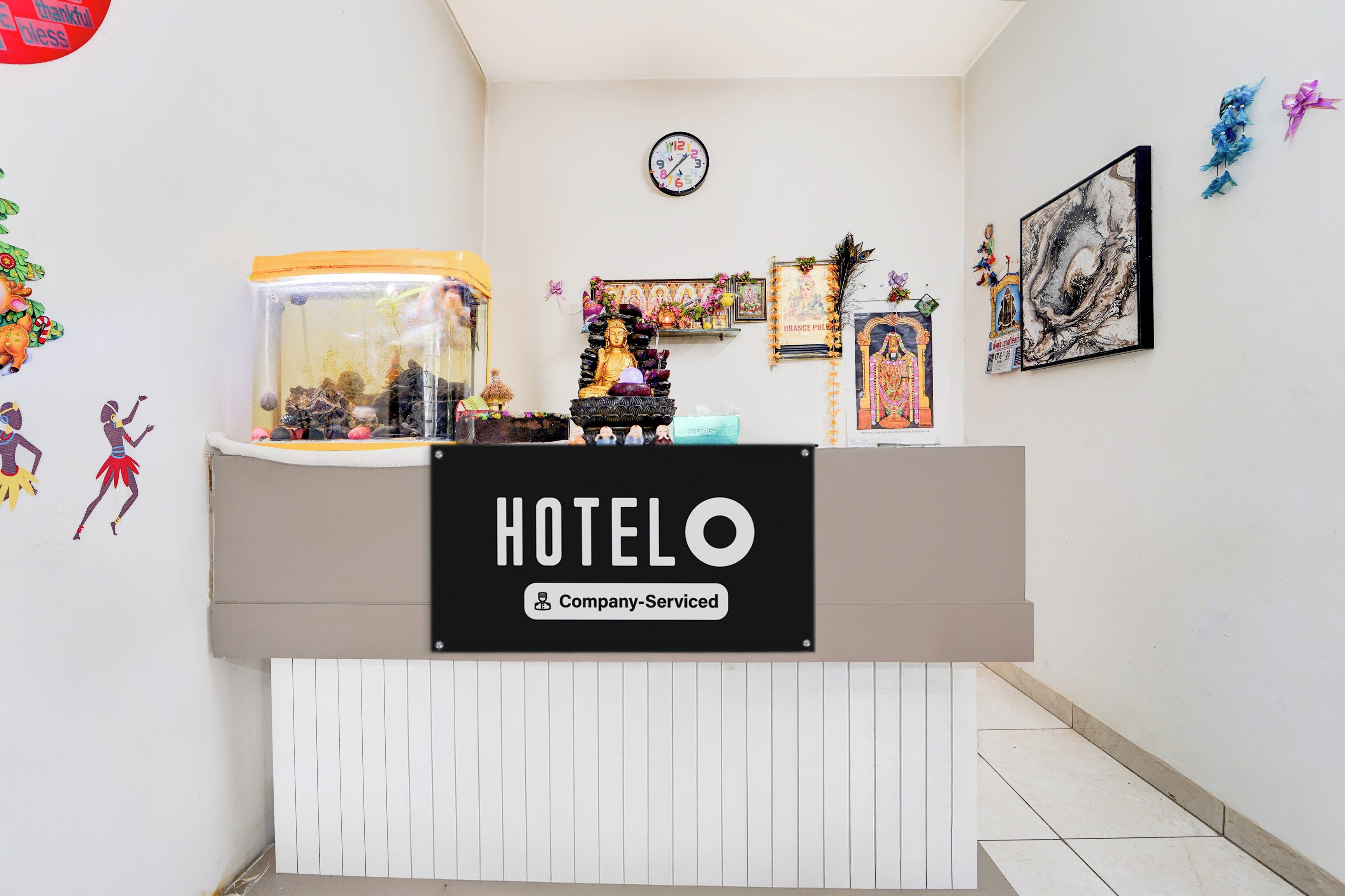 A hotel reception desk with a clock above it, a sign indicating company service, and various decorations including a peacock feather and framed pictures on the wall.