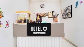 A hotel reception desk with a clock above it, a sign indicating company service, and various decorations including a peacock feather and framed pictures on the wall.