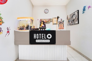 Un mostrador de recepción de hotel con un reloj encima, un cartel que indica servicio de empresa y varias decoraciones, incluyendo una pluma de pavo real e imágenes enmarcadas en la pared.