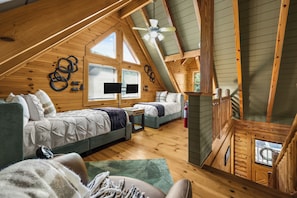 Ein gemütliches Schlafzimmer im Dachgeschoss mit schrägen Wänden, Holzbalken, zwei Betten, einem Deckenventilator und einem Fenster mit Jalousien.