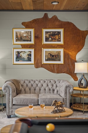 A room with a large wall-mounted cowhide, a tufted sofa, a round coffee table with glasses, and framed pictures. - Private Indoor Pool | Arcade Loft & Hot Tub (Sevierville)