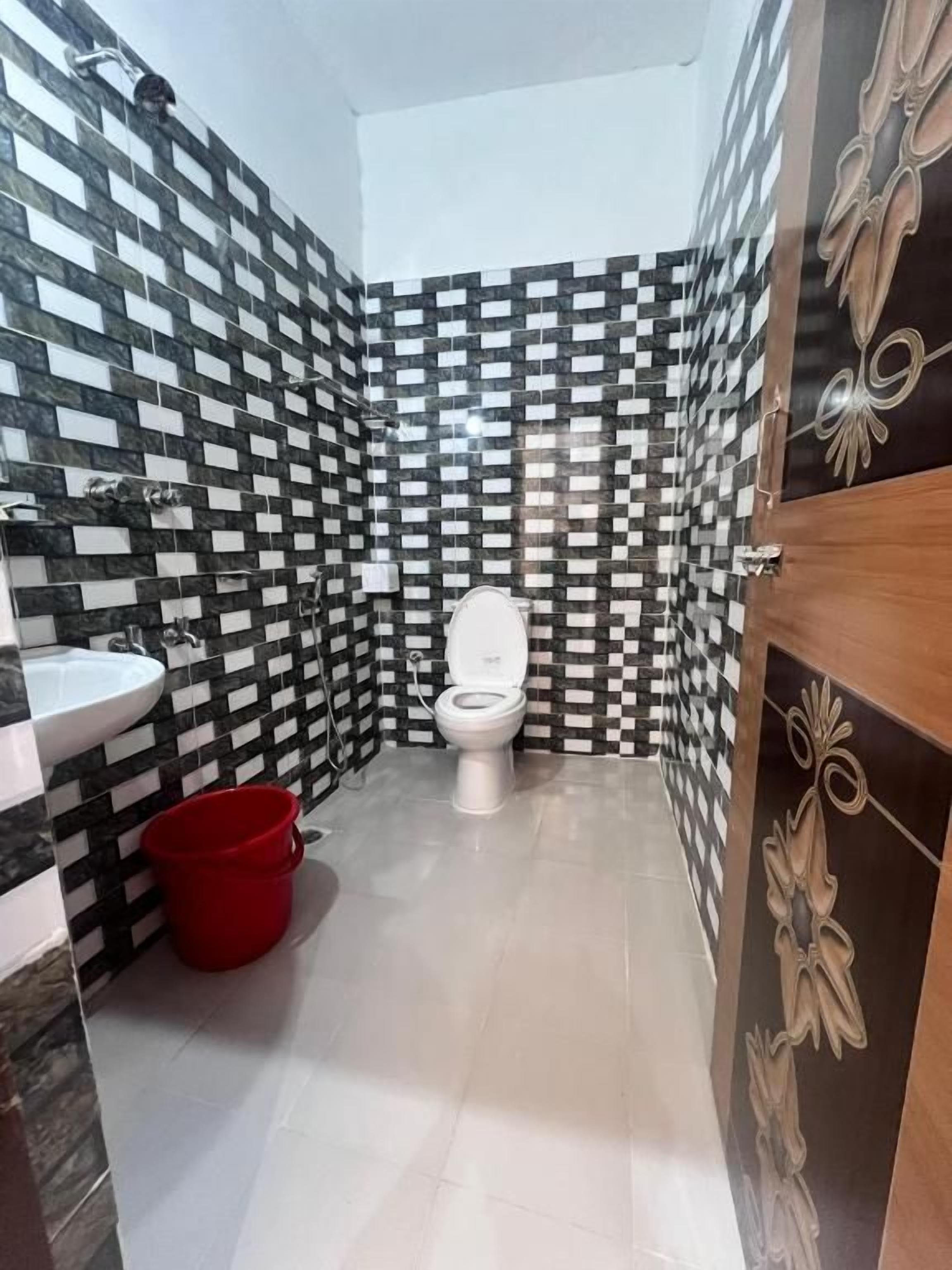 Un baño con pared de azulejos blancos y negros a cuadros, un inodoro blanco, un cubo rojo y una puerta de madera con un tirador decorativo.