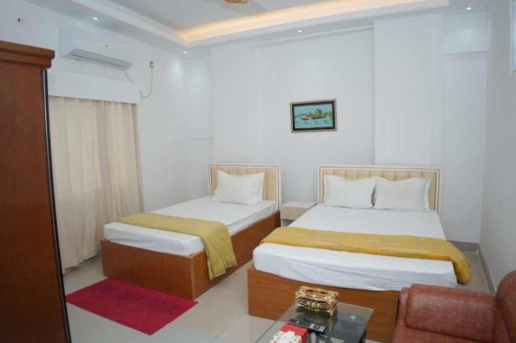 A hotel room with two single beds, a small table, a chair, and a red rug.