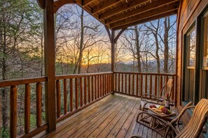 Une terrasse en bois offrant une vue sur les arbres et les montagnes, une table avec de la nourriture et des boissons, et une chaise.