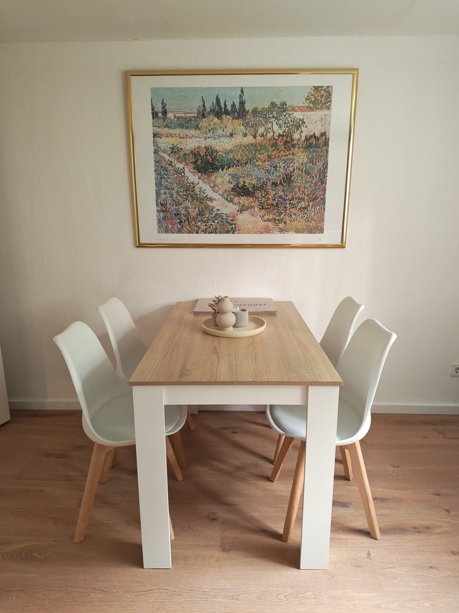 A dining area with a wooden table and white chairs, a framed painting on the wall, and a small vase on the table.