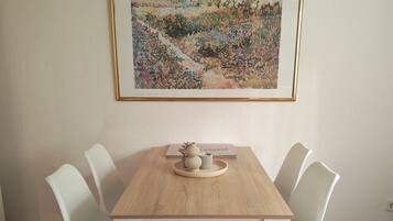 A dining area with a wooden table and white chairs, a framed painting on the wall, and a small vase on the table.