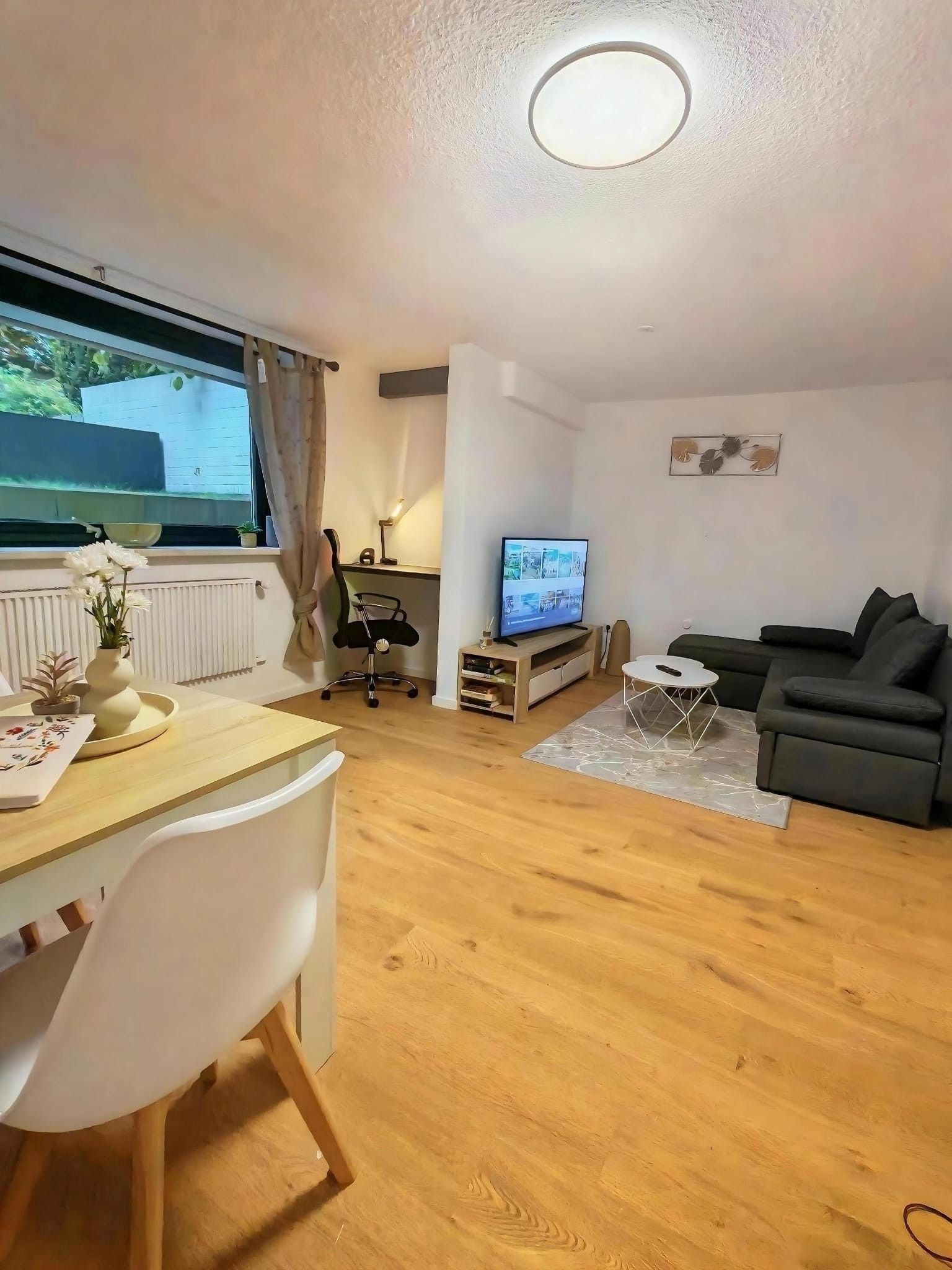 A modern living room with a wooden floor, a dining area with a white table and chairs, a black office chair, a television, and a black sofa.