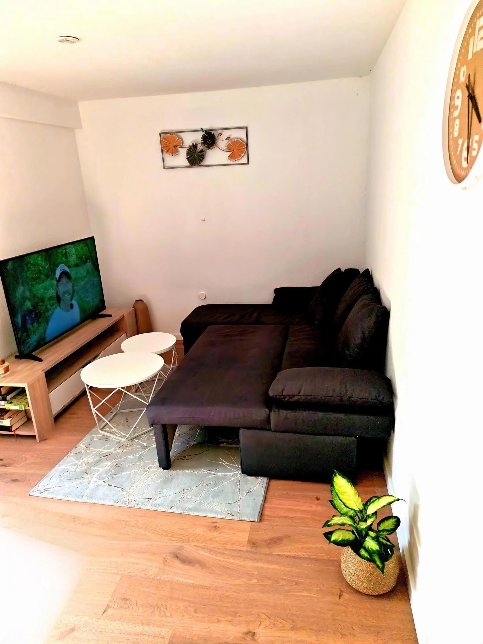Ein Wohnzimmer mit einem dunkelbraunen Sofa, einem kleinen weißen Tisch, einem Fernseher und einem Wandbild.