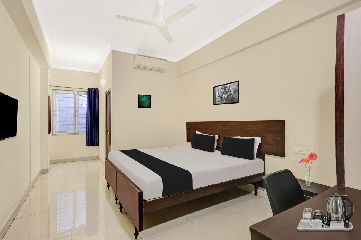 A hotel room with a bed, a desk with a kettle, a chair, a window with curtains, and a ceiling fan.