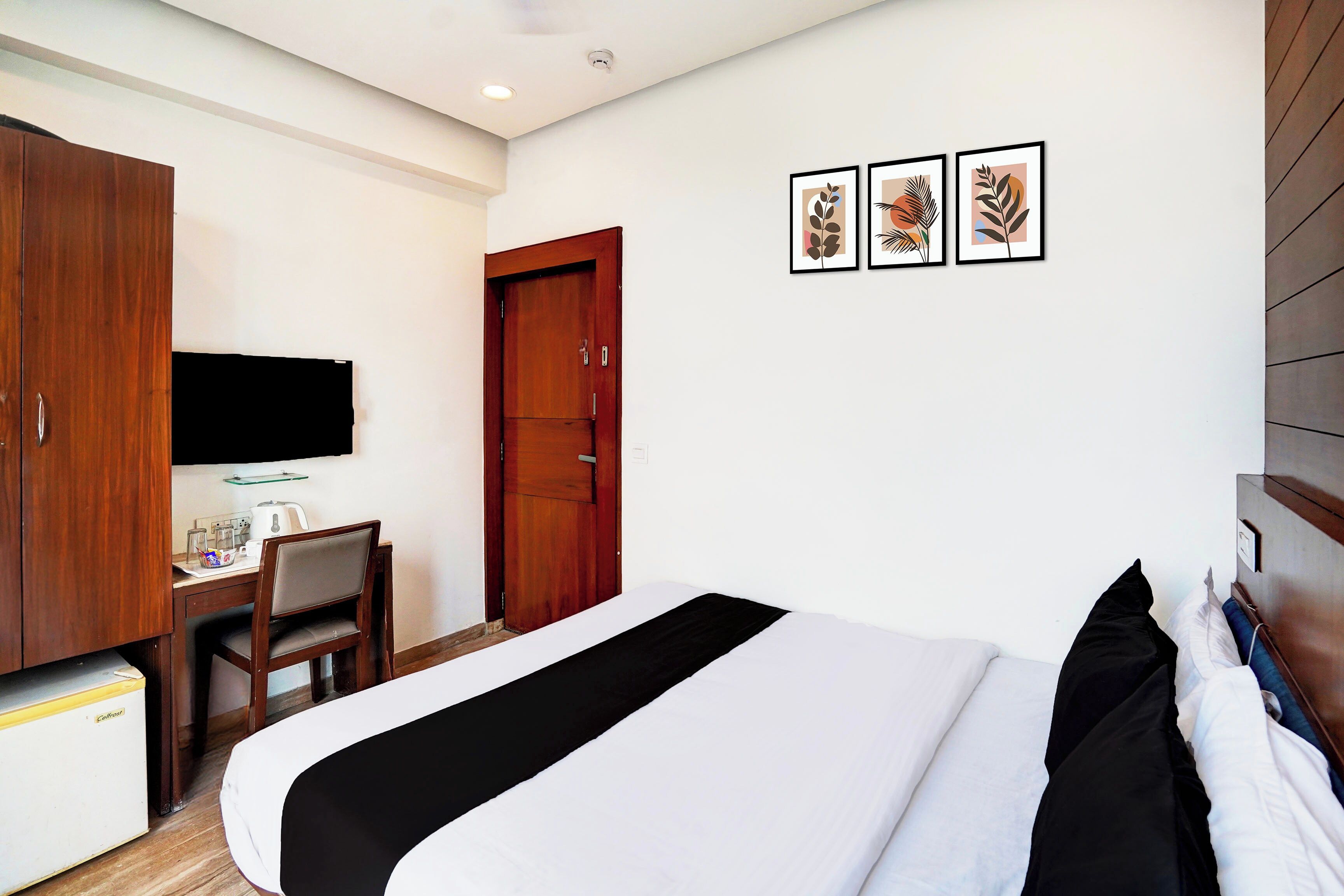 A hotel room with a bed, a desk with a chair, a wardrobe, and three framed pictures on the wall.
