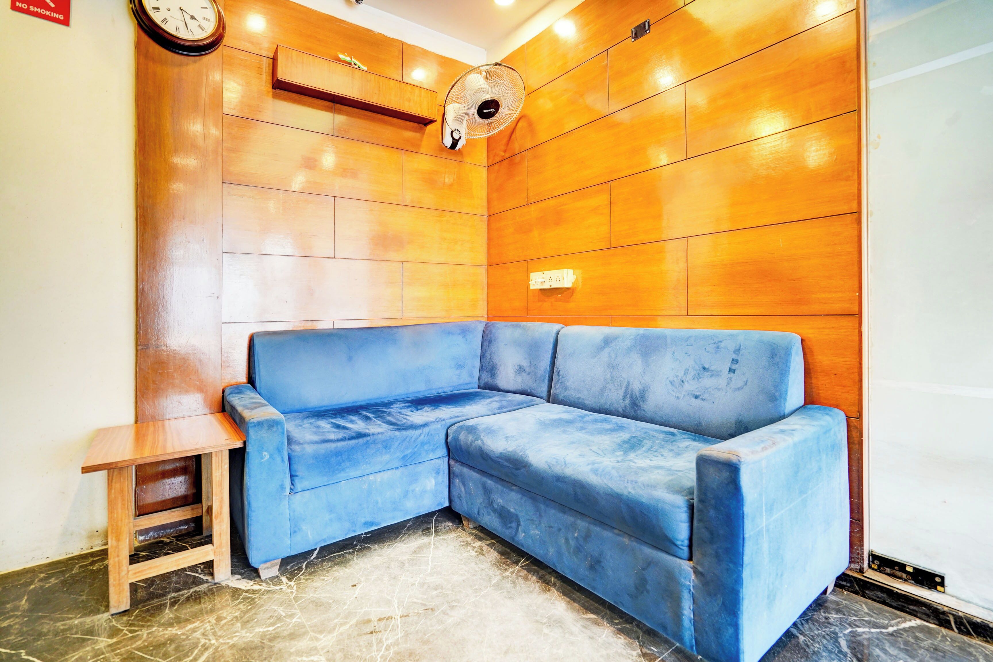A corner seating area with a blue sofa, a small wooden table, and a wall-mounted fan.