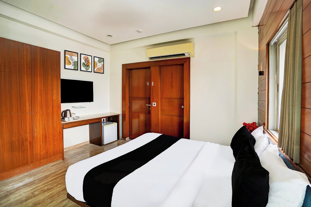 A hotel room with a bed, a desk with a flat-screen TV, a wardrobe, and an air conditioning unit.