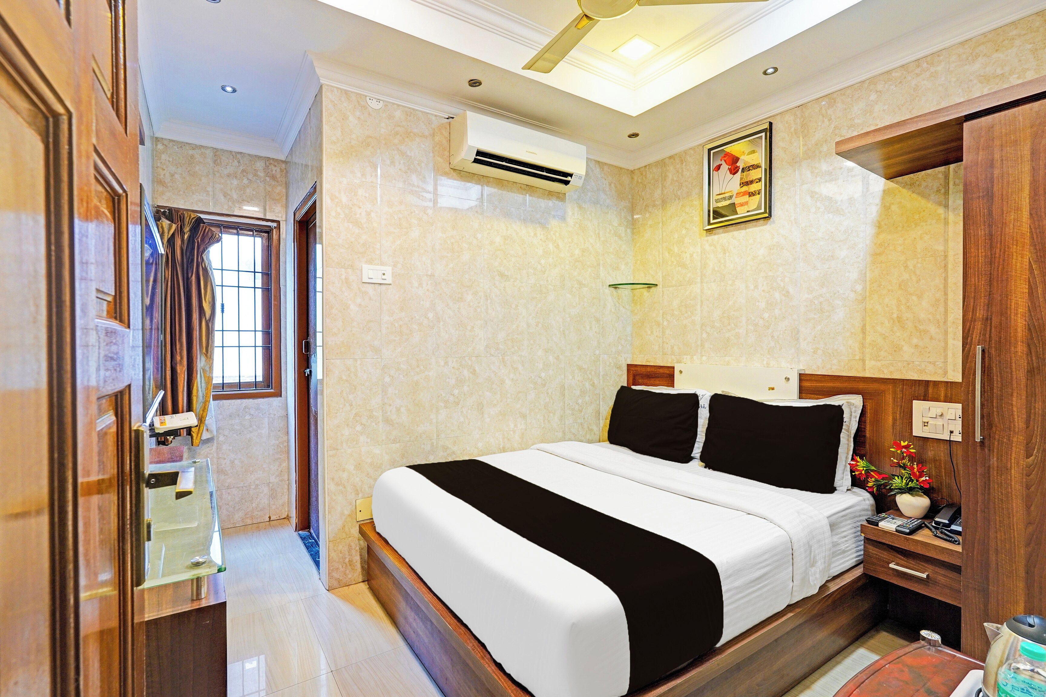 A hotel room with a bed, bedside table, nightstand, mirror, and an air conditioning unit.