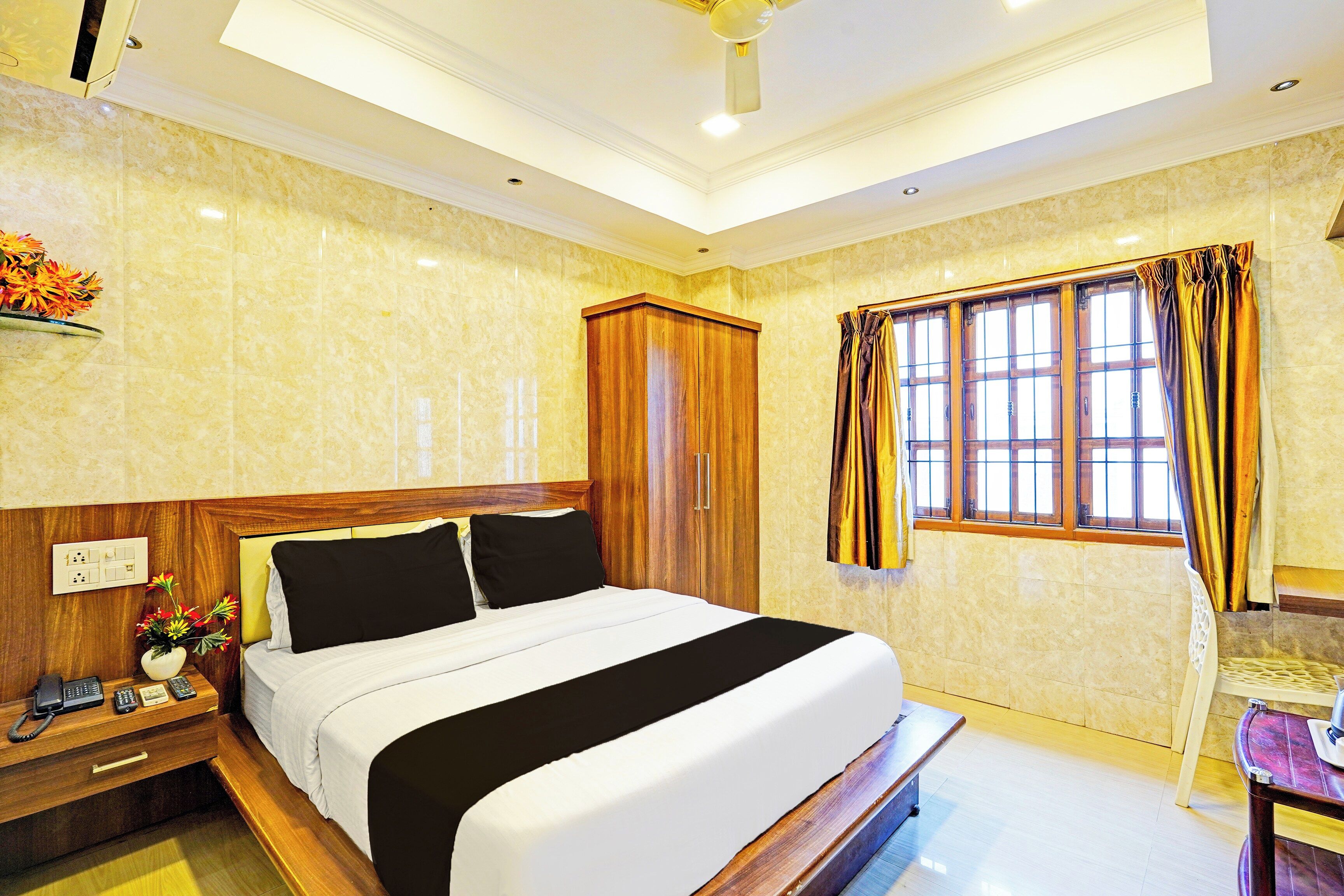 A hotel room with a bed, bedside table, a telephone, a vase with flowers, a window with curtains, and a ceiling fan.