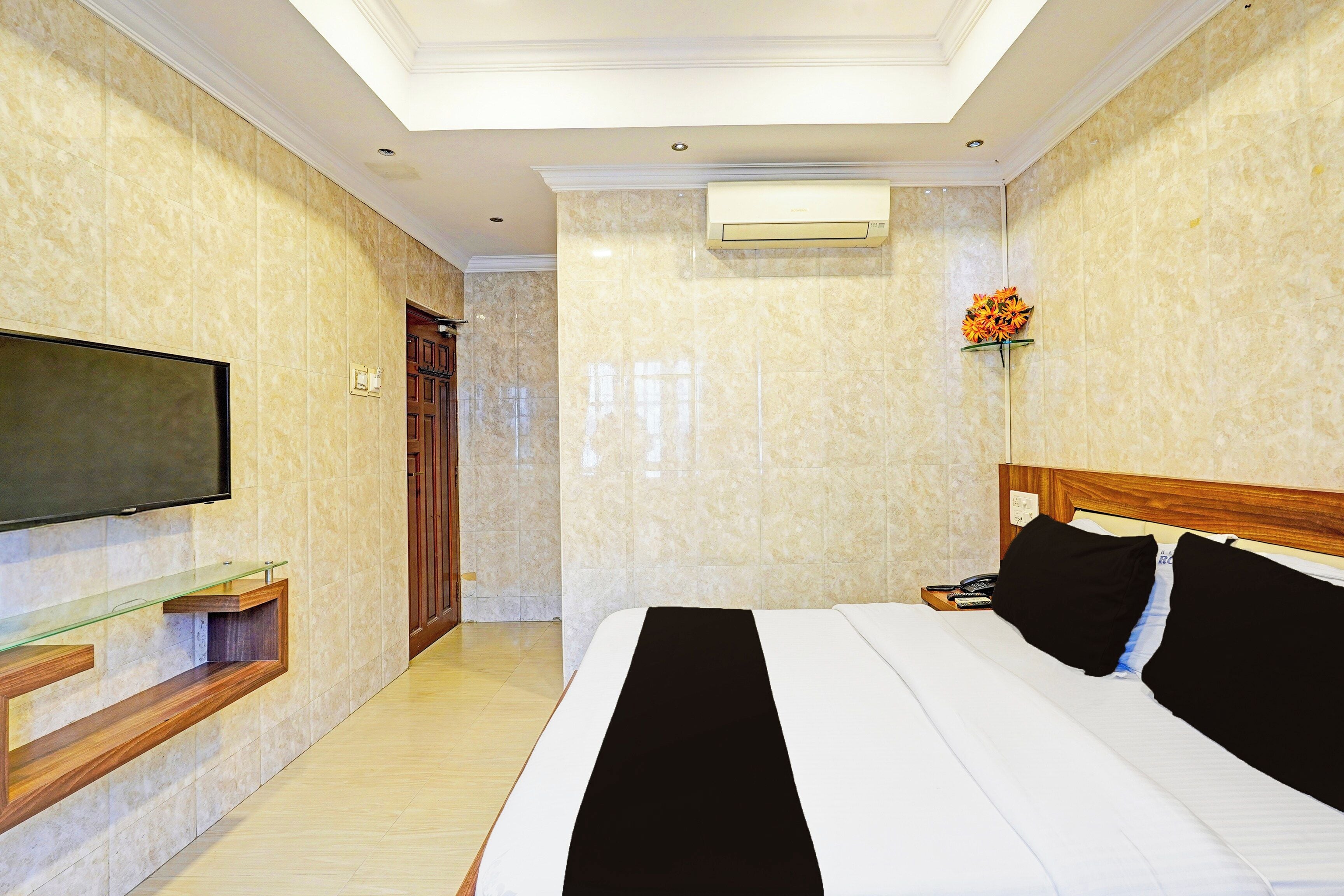 A hotel room with a bed, a television mounted on the wall, a wooden nightstand, and a door leading to another room.