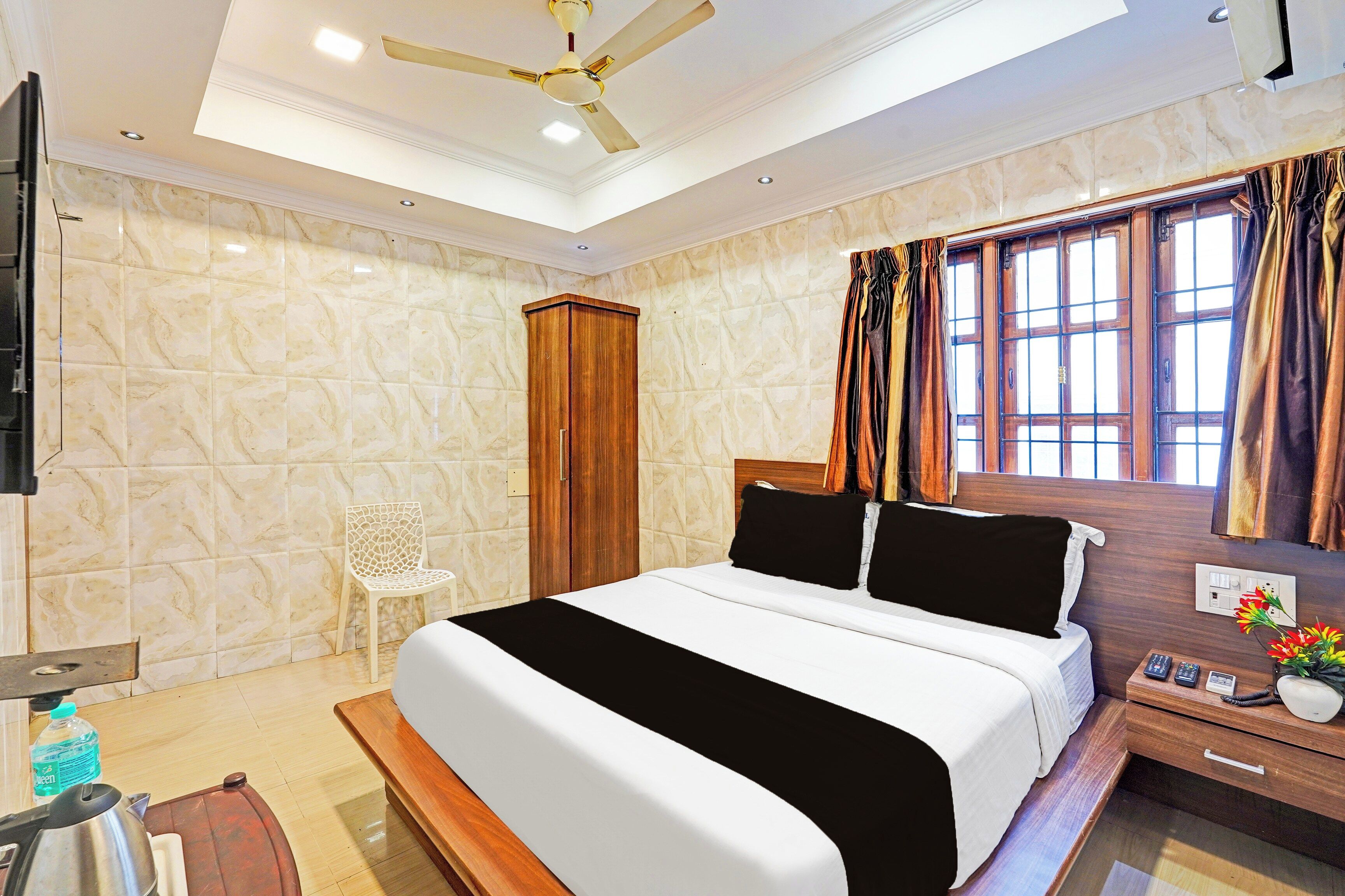 A hotel room with a bed, a wooden wardrobe, a chair, a bedside table with a phone and a vase, and a window with curtains.