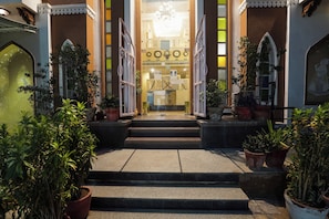 Entrada de hotel con escalones que conducen a una puerta de vidrio, flanqueada por macetas y detalles decorativos.
