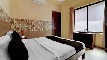 A hotel room with a bed, a window with curtains, a bedside table with a vase of flowers, and a wall-mounted air conditioner.