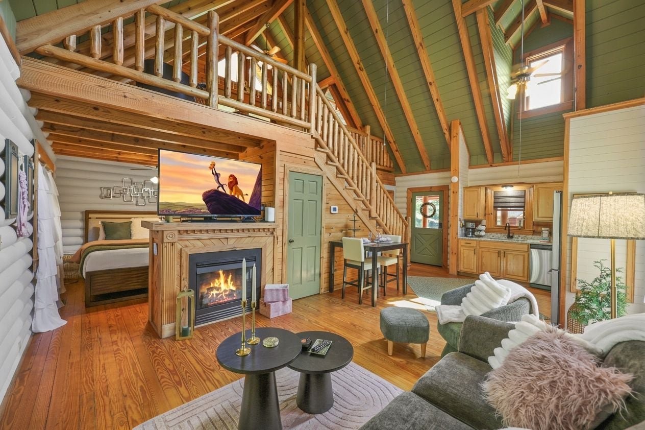 A log cabin interior with a fireplace, a dining area, a kitchen, and a loft bedroom.