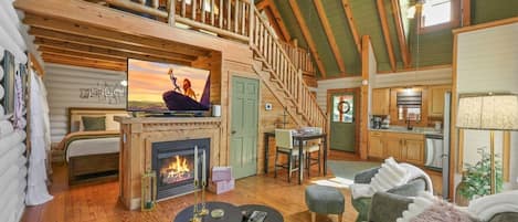 A log cabin interior with a fireplace, a dining area, a kitchen, and a loft bedroom.