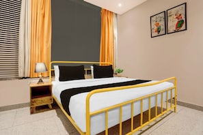Ein ordentlich bezogenes Bett mit gelbem Gestell, weißer Bettwäsche und schwarzen Kissen. Ein Nachttisch mit Lampe, einer Topfpflanze und einem gerahmten Bild an der Wand.