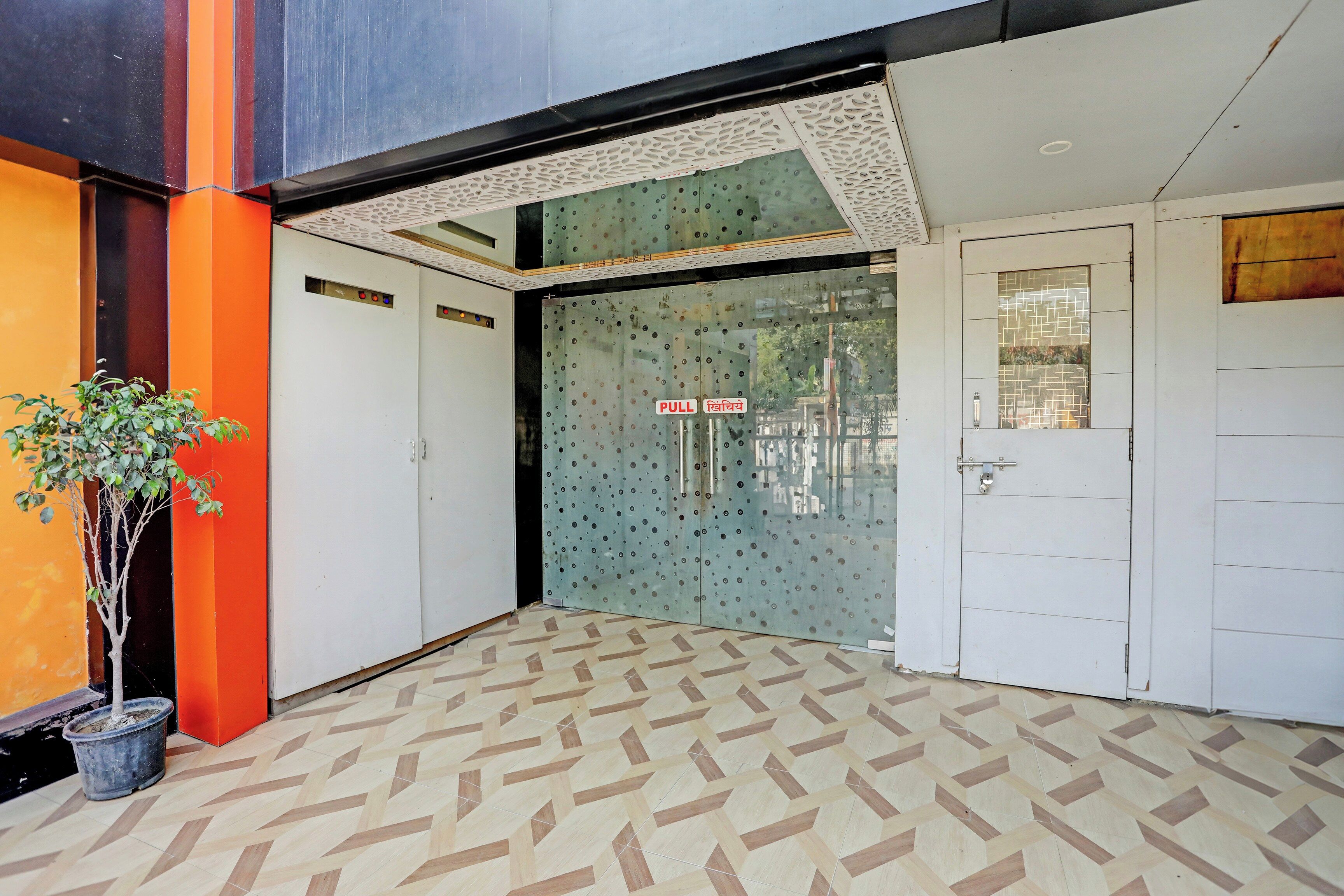 A hotel entrance with a geometric patterned floor, a potted plant, and a glass door with a 'PULL' sign.