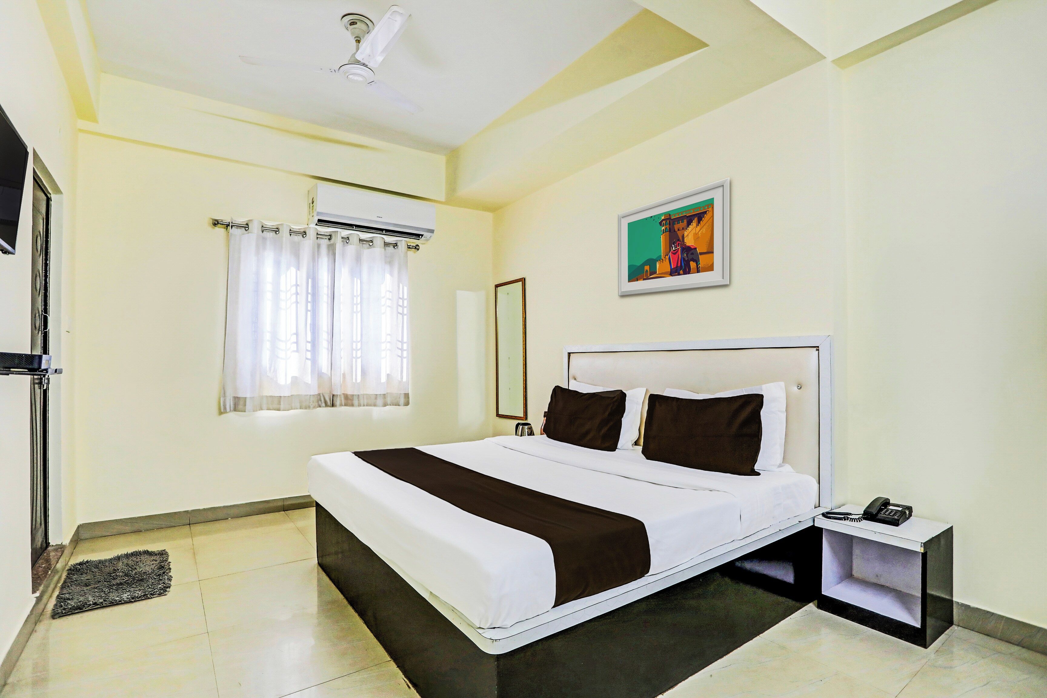 A hotel room with a bed, a nightstand, a telephone, an air conditioning unit, a window with curtains, and a painting on the wall.