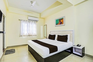 Una habitación de hotel con cama, mesita de noche, teléfono, equipo de aire acondicionado, ventana con cortinas y un cuadro en la pared.